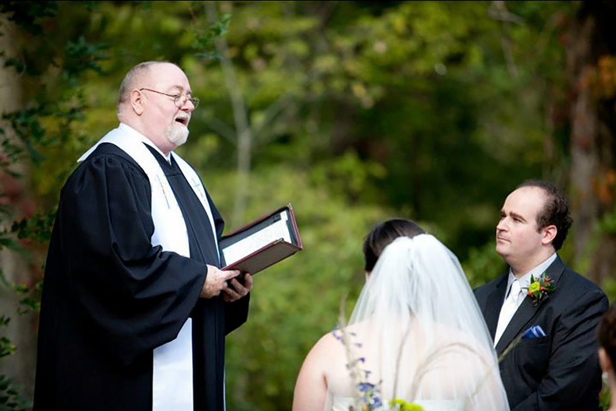 The Marrying Rev reciting wedding vows to a bride and groom at an outdoor wedding